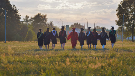 Recent graduates celebrating their academic success and friendship as they walk through a field at sunset, holding handsの写真素材