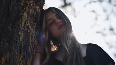Portrait of a teenage girl with long hair leaning against a tree in a forest, enjoying the sunlight...
