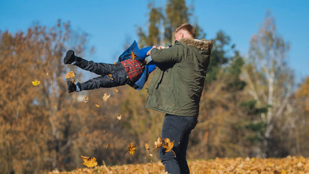 Father sharing playful interaction with son amid golden autumn leaves, bright sunlight filtering through park landscapeの写真素材