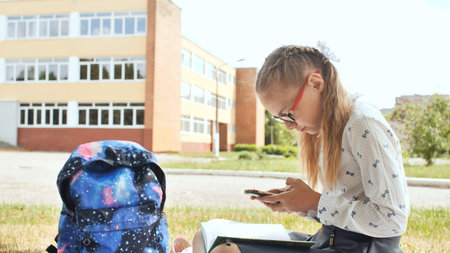 Schoolgirl using smartphone sitting on grass near school building after class, with backpack and...