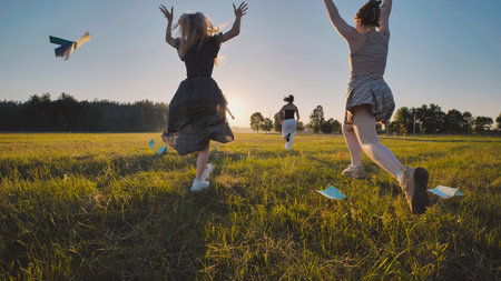 High school girls having fun running in a field at sunset, tossing paper airplanes during their...