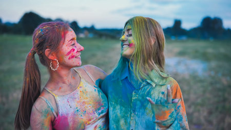 Two vibrant young women, covered in holi powder, share a joyful embrace at a colorful festival...