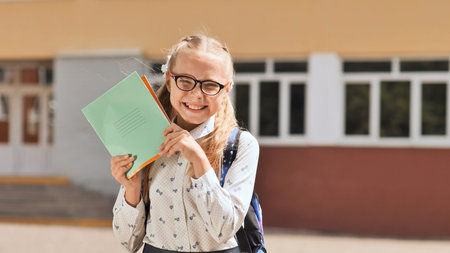 Happy schoolgirl holding notebooks and smiling in front of school building, ready for studying and learning new thingsの写真素材