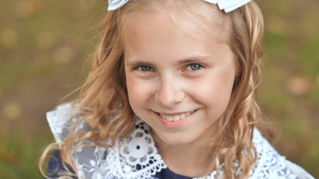Close up portrait of a smiling schoolgirl with white bows in her blond hair, enjoying her time outdoorsの写真素材