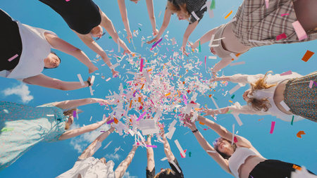 High school graduates joyfully tossing colorful confetti into the blue sky to celebrate their...