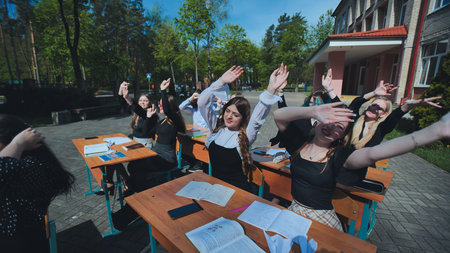 Happy students raising hands sitting at desks outdoors, celebrating the end of the school yearの写真素材