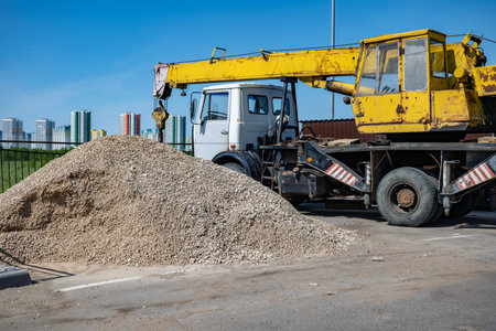 Mobile crane operating at construction site with pile of gravel, city buildings background