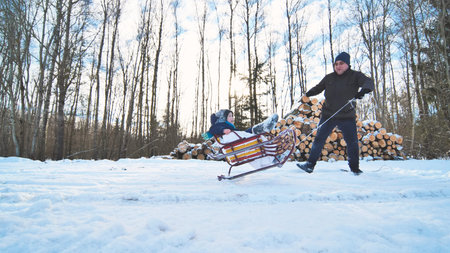 Dad pulls his son on a sled in a snowy forest next to a pile of firewood on a sunny winter day
