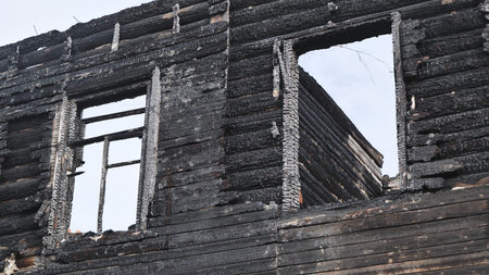 Charred ruins of a burnt wooden log house with empty window frames against an overcast sky.