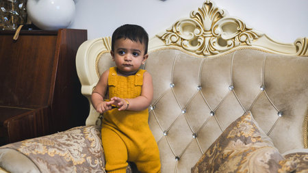 Cute baby boy in yellow romper standing on a sofa at home