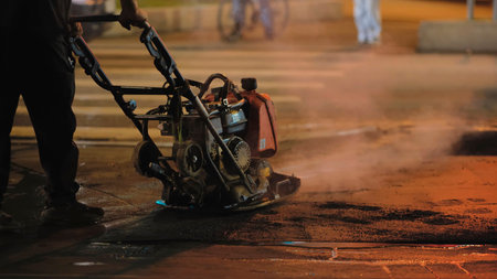 Road worker using vibrating plate compactor on fresh asphalt at night city street