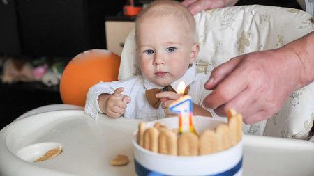 Baby boy celebrating first birthday with cake, candle, and helping hand during party