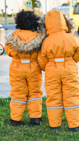 Two young kids in warm padded winter clothing standing on grass, looking forward