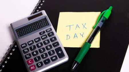 Tax day written on a yellow sticky note next to a calculator and a pen.の写真素材
