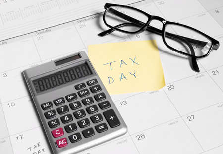 Tax day written on a sticky note next to a calculator and glassesの写真素材
