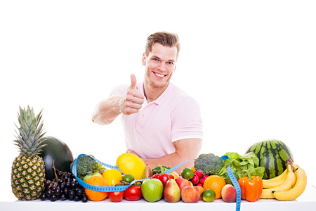 He knows what's good for you! - Handsome muscular man sitting behind a table full of vegetables and fruits and showing thumb up, isolated on whiteの写真素材