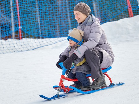 In winter, happy mom and son (3 years old) ride a snow scooter from the mountain in pure white snow.の写真素材
