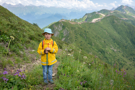 A boy traveler with trekking poles in a yellow raincoat and a panama is standing on the path to the top of the mountain range. Caucasus, Russiaの写真素材