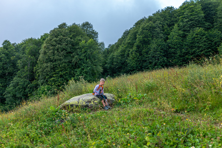 A boy traveler with a backpack and trekking poles is resting on a stone on the mountainside.の写真素材