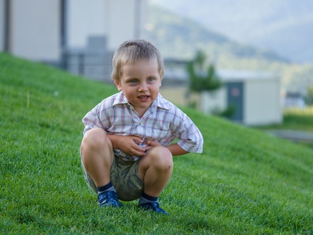 A little boy sits on a green slope against the backdrop of mountainsの写真素材