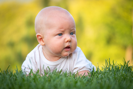The baby lies or crawls along the green lawn at sunset and looks indignantly. Emotions baby. Vacation with children. Childcare. Goods for kids. Sun protection. Child development.の写真素材