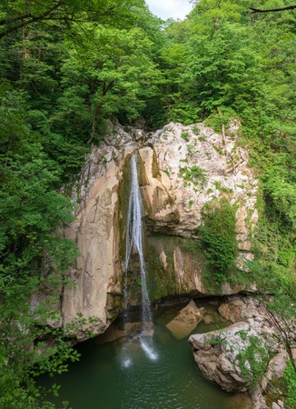 A high waterfall falls from a cliff into a clear lake. The waterfall is surrounded by green forest. River Agura, Sochi, Russiaの写真素材
