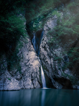 A tall waterfall falls from a cliff into a clear lake at night, lit by the light of the moon. The waterfall is surrounded by green forest. River Agura, Sochi, Russiaの写真素材