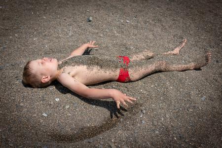The boy lies on the beach on a sunny day and is covered with small pebbles.の写真素材