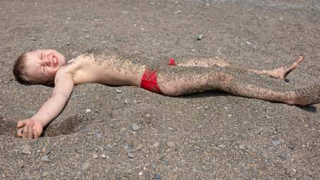 The boy lies on the beach on a sunny day and is covered with small pebbles.の写真素材