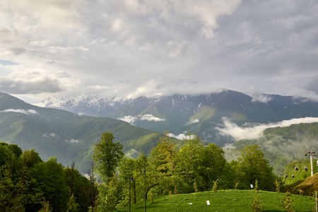 Green trees and cable car on the background of high snowy mountains. Spring colors. Cloudy spring day in the mountainsの写真素材