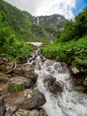 Views of the green mountains with rocks and the highest waterfall. Powerful stream of a mountain river among stones and rocks. Unmelted snow in the high mountainsの写真素材