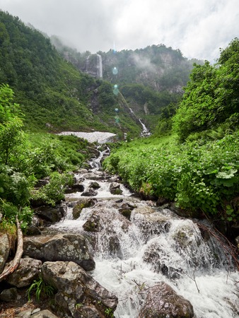 Views of the green mountains with rocks and the highest waterfall. Powerful stream of a mountain river among stones and rocks. Unmelted snow in the high mountainsの写真素材
