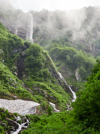 Views of the green mountains with rocks and the highest waterfall in a dense fog. Powerful stream of a mountain river among stones and rocks. Unmelted snow in the high mountainsの写真素材