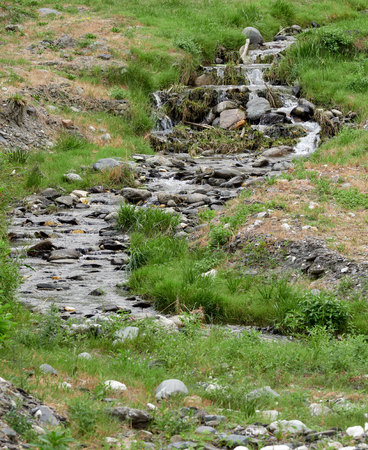 A stony stream flows through a green meadow with low grass. A small waterfall on the creek.の写真素材
