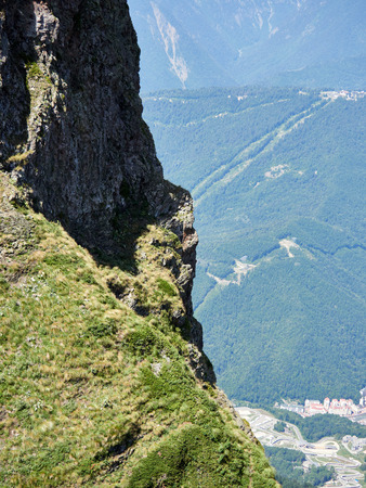 The rock on a high mountain slope, in front of which is a valley with residential houses. Blue sky in the mountains on a summer day.の写真素材
