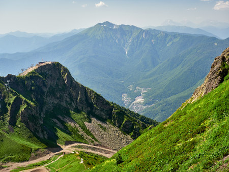The peaks of a high mountain slope with cable car, in front of which is a valley with residential houses. Blue sky in the mountains on a summer day. Krasnaya Polyana, Sochi, Russiaの写真素材