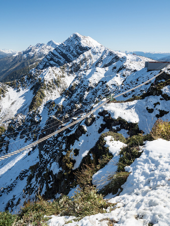Suspended pedestrian bridge in the high snowy mountains. Clear blue sky. Mountains with green slopes.の写真素材
