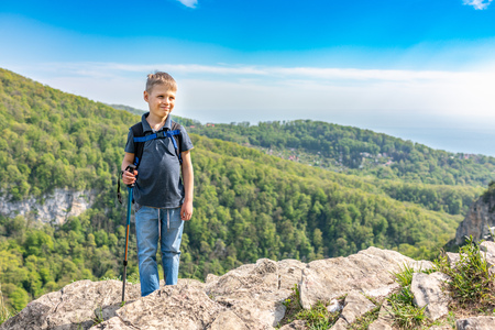 A smiling boy traveler with trekking poles and a backpack stands on top of a mountain among a green forest. Clear spring or summer day in the mountainsの写真素材