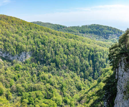 Mountain with a steep rocky slope and valley with thick green forest below. Residential buildings and the sea are visible in the distanceの写真素材