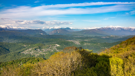 View at sunset on a green hilly valley with high snowy mountains on the horizon. Green valley with village settlements in the light of sunset. View from Mount Akhun, Sochi, Russia.の写真素材