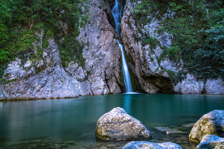 A high waterfall falls from a cliff into a clear lake. The waterfall is surrounded by green grass and forest. River Agura, Sochi, Russiaの写真素材
