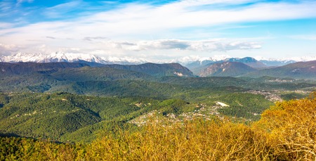 View at sunset on a green hilly valley with high snowy mountains on the horizon. Green valley with village settlements in the light of sunset. View from Mount Akhun, Sochi, Russia.の写真素材