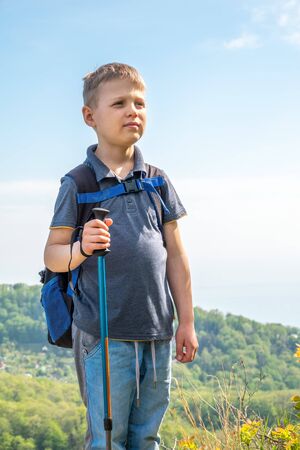 A boy traveler with trekking poles and a backpack stands on top of a mountain among green forest. Clear spring or summer day in the mountainsの写真素材