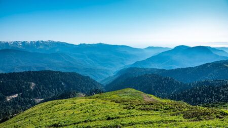 The view from the height of a green mountain valley surrounded by high mountains. Snow-capped mountain peaks on the horizon. Krasnaya Polyana, Sochi, Caucasus, Russia.の写真素材