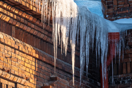 Large icicles hanging from the roof of an old brick building. Roof of old building covered with sharp icicles. Ice stalactite. Poor thermal insulation of the roof leads to the formation of iciclesの写真素材