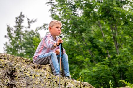 A boy traveler 7 years old with a backpack and trekking poles is resting on a stoneの写真素材