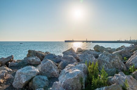 Large stones and rocks against the background of the seaport. Breakwater of large stones on the sea coast. Clear sky and bright sunset at sea port with a breakwater and a lighthouseの写真素材