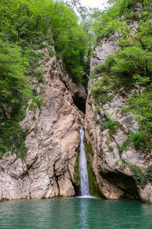 A high waterfall falls from a cliff into a clear lake. The waterfall is surrounded by green forest. River Agura, Sochi, Russiaの写真素材