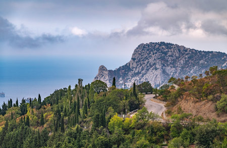 Rocky mountain on sea shore with high green trees on slope. Seascape with cloudy sky. Old cemetery...