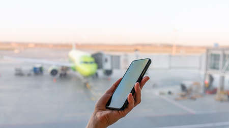 Close-up shot of female hand holding smart phone by window at airport. Area with airplane in background. Departure for vacation, waiting for the plane at the airport. Flight delay.の写真素材